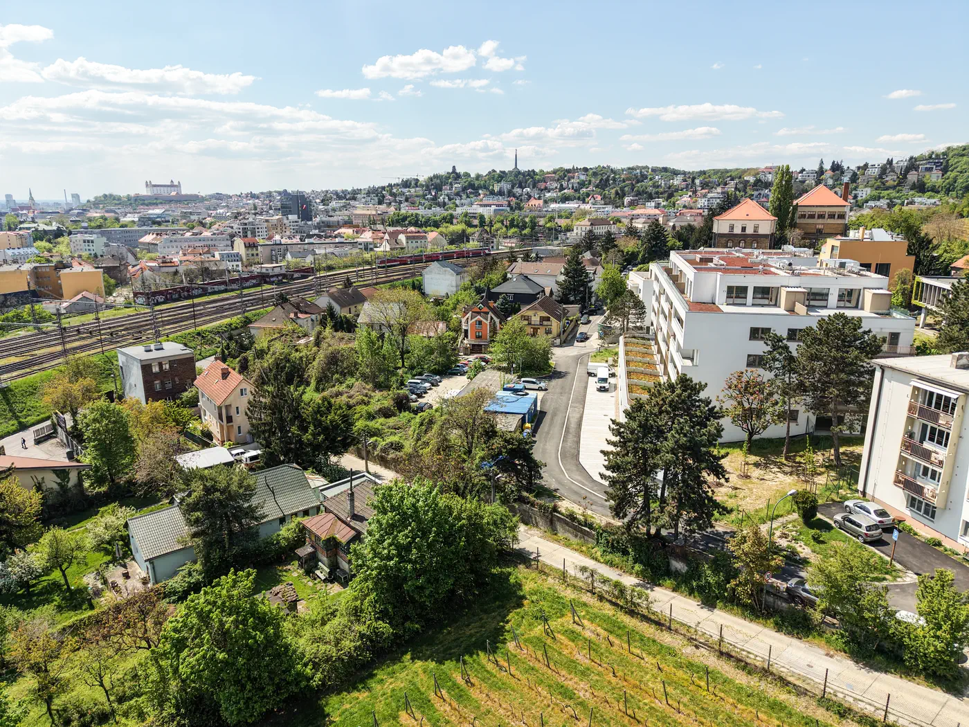 Pohľad z dronu na bytový dom Winehills, vinice nad ním a mesto s hradom na obzore